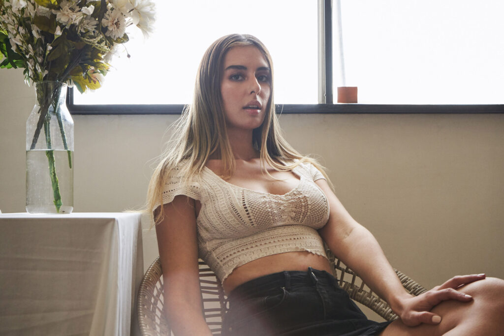 Marintia Bustos a model with long blonde hair sits in a wicker chair by a window wearing a white lace top and black skirt. Beside her is a vase with white flowers.
